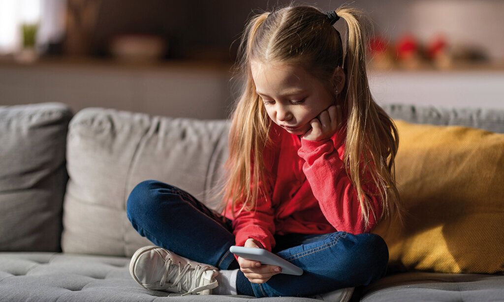 Kleines Mädchen  sitzt im Schneidersitz auf dem Sofa und daddelt am Smartphone, guckt gelangweilt