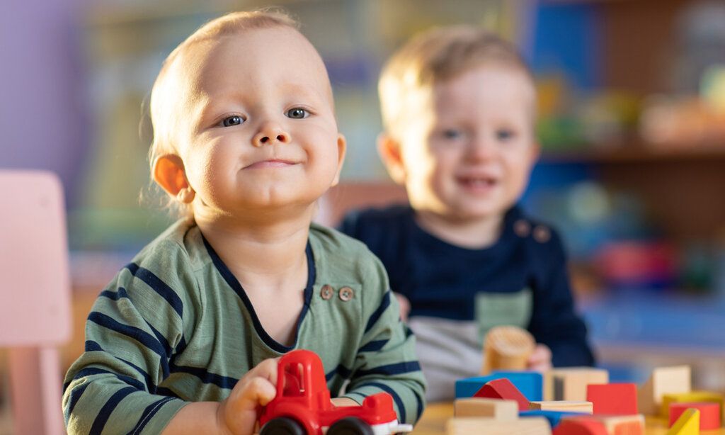 zwei Kleinkinder spielen in einer Kita mit Bauklötzen und Spielzeugauto, ein Kind ist im Vordergrund, das andere im Hintergrund unscharf