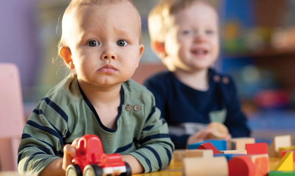Kleinkind spielt mit Holzspielzeug und guckt skeptisch in die Kamera, im Hintergrund ein lachendes Kleinkind, unscharf