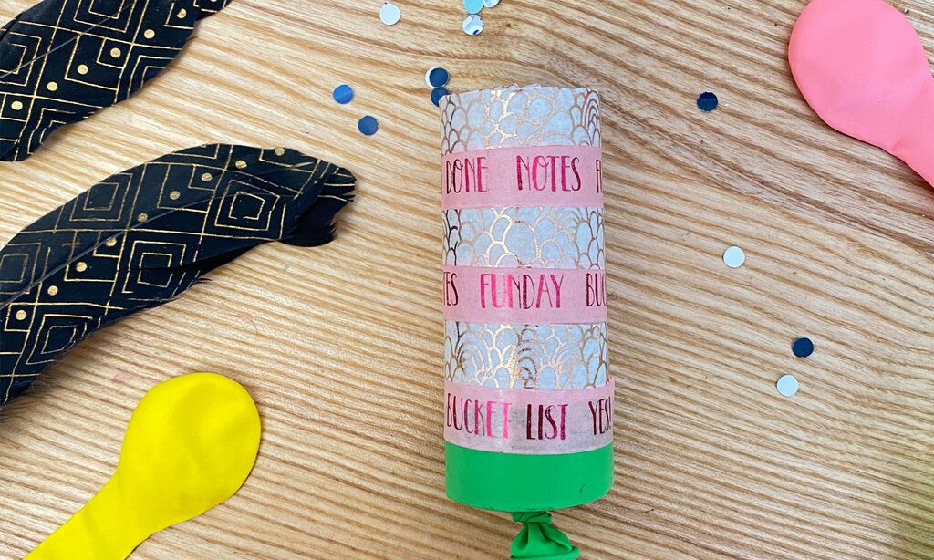 selbstgebastelte Konfettikanone auf Holztisch, daneben Federn, Luftballons und Pailletten