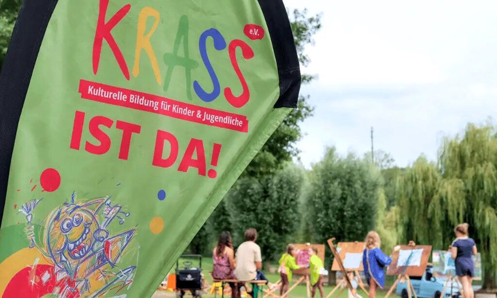 Beachflag mit Krass-Logo im Vordergrund, im Hintergrund Kinder, die an Staffeleien malen