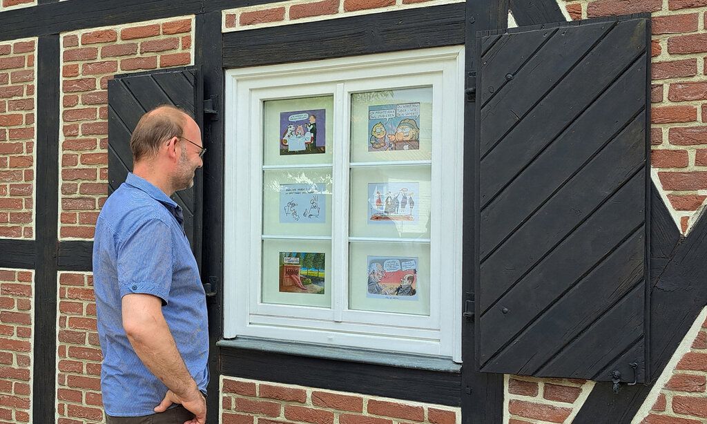 Ein Mann steht vor dem Fenster eines Fachwerkhauses, in dem Cartoons präsentiert werden, und grinst