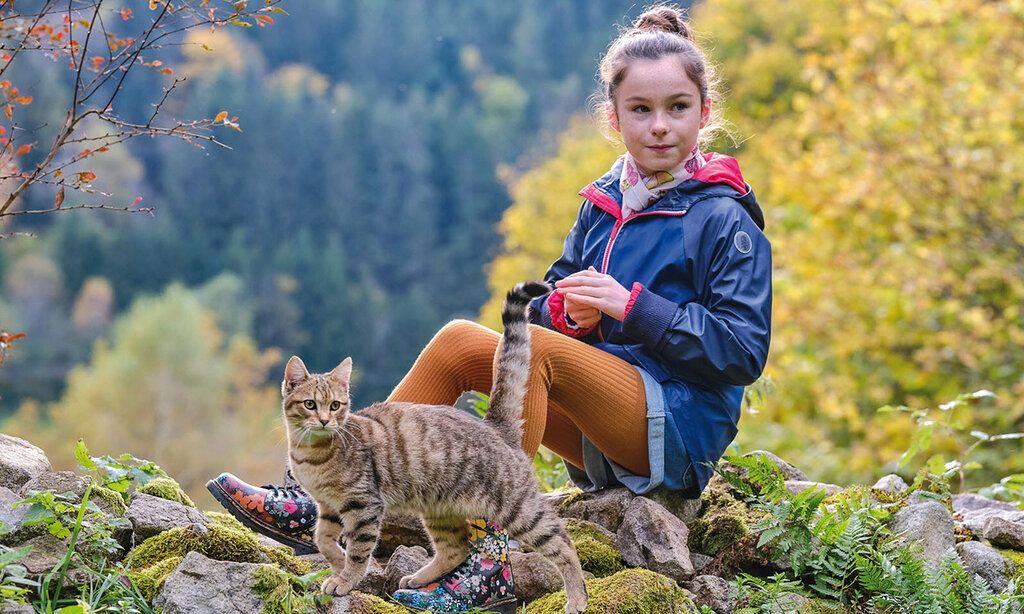 Ein Mädchen sitzt in der Natur auf einem Stein, im Hintergrund Wald und im Vordergrund eine Katze