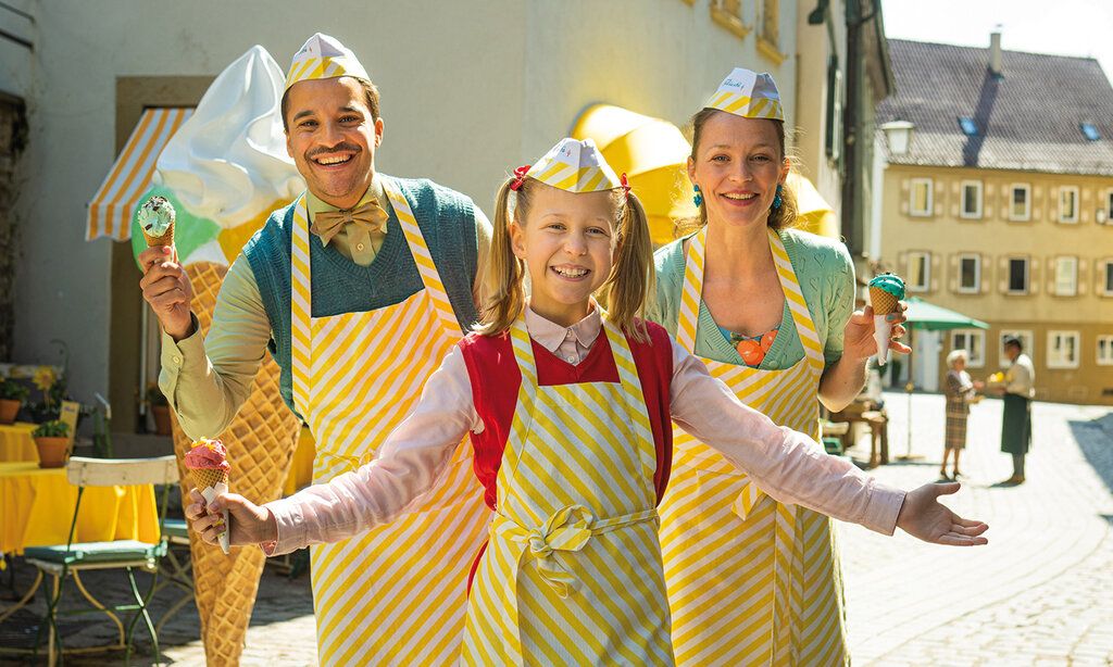 Ein Mädchen steht mit Vater und Mutter auf einem Platz, alle drei tragen Eisverkäufer-Schürzen und -hütchen und ein Eis im Hörnchen