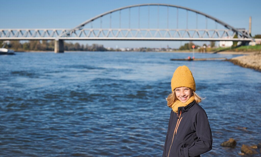 Mädchen mit Mütze steht am Rhein, im Hintergrund die Eisenbahnbrücke von Düsseldorf-Hamm