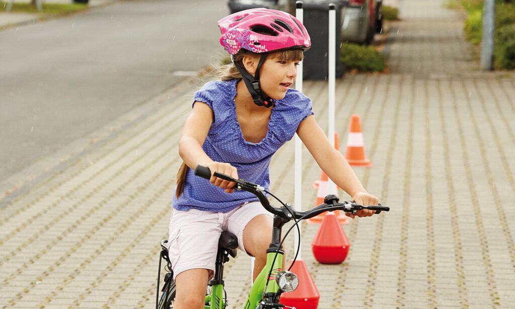 Ein Mädchen mit einem pinken Fahrradhelm fährt mit dem Fahrrad einen Parcours, der mit Verkehrshütchen abgesteckt ist
