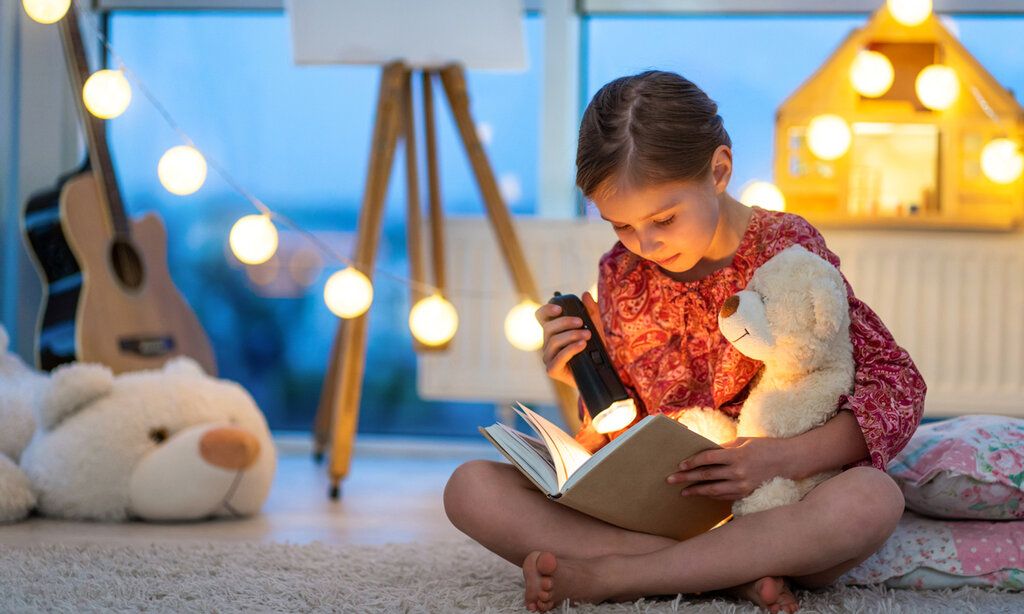 Mädchen sitzt mit einem Teddy auf dem Boden und liest mit einer Taschenlampe in einem Buch, weihnachtliche Stimmung