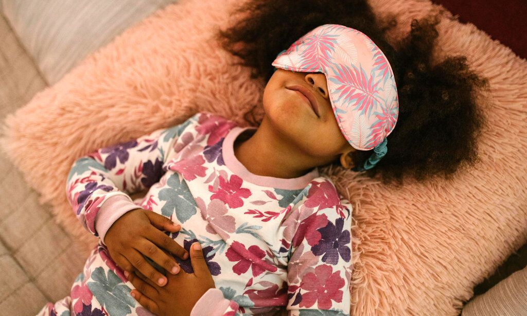 ein Mädchen liegt mit einem geblümten Schlafanzug und einer geblümten Schlafmaske auf einem plüschigen Kissen