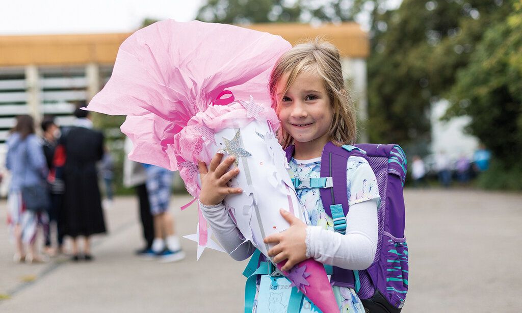 Mädchen steht mit einer großen Schultüte auf dem Schulhof, im Hintergrund eine Gruppe von Leuten