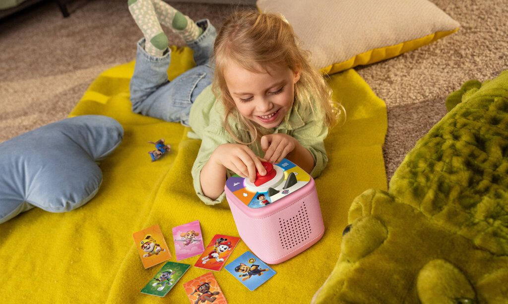 Mädchen liegt mit Toniesbox und Spielkarten auf einer Decke auf dem Boden