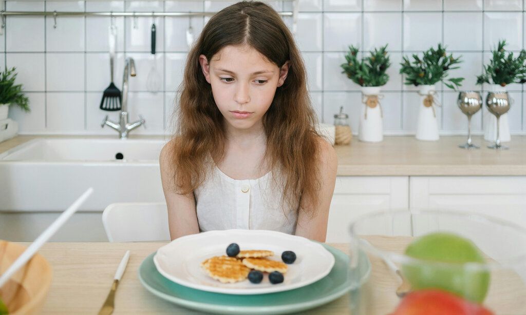 Mädchen sitzt am Küchentisch, vor sich ein Teller mit Eaffeln und Blaubeeren und  möchte nicht essen