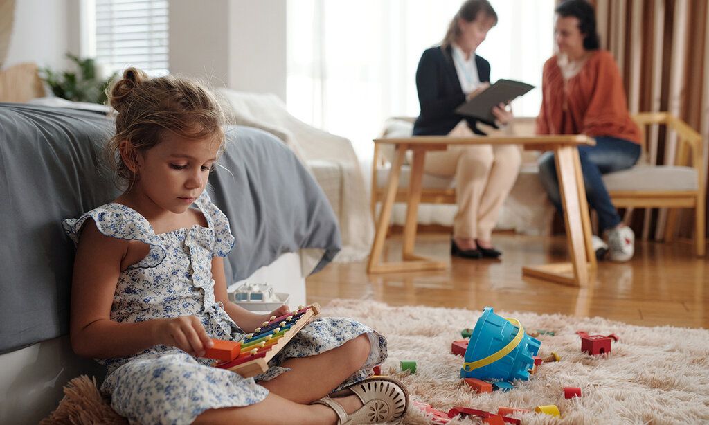 Mädchen sitzt im Wohnzimmer und spielt, zwei Frauen sitzen im Hintergrund