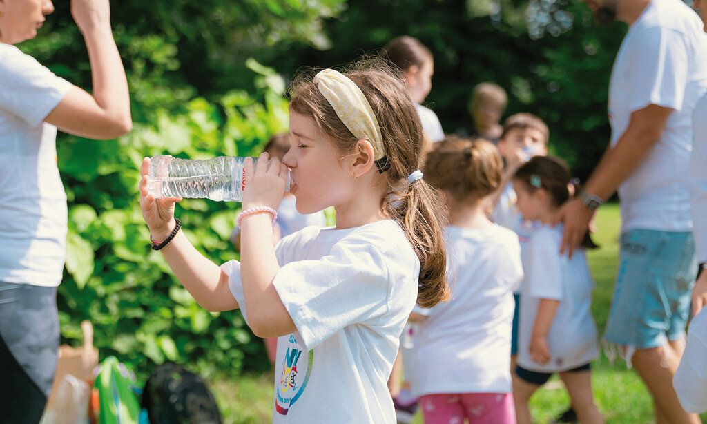 Mädchen trinkt aus Wasserflasche, im Hintergrund andere Kinder und Erwachsene ineinem Park