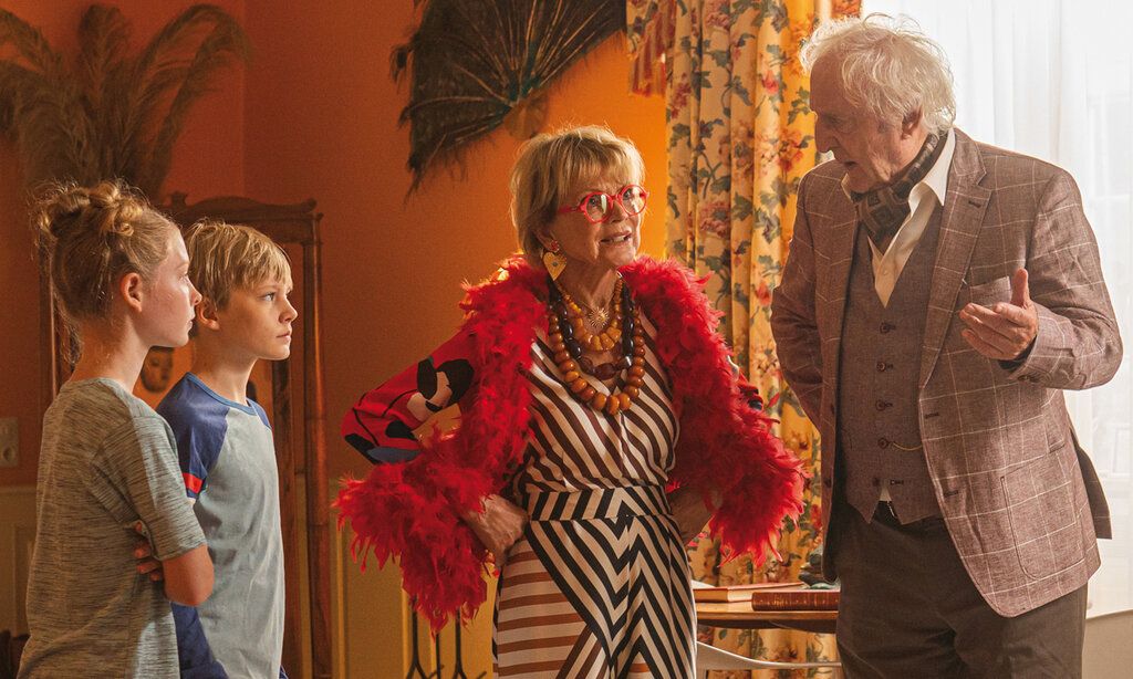 Ein Junge und ein Mädchen steh mit einer älteren Dame und einem älteren Herrn zusmmen in einem Wohnzimmer, die Dame trägt eine rote Feder-Boa, Klunker und eine rote Brille