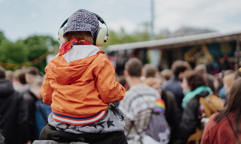 ein Kind mit Kopfhörern auf den Schultern eines Erwachsenen, von hinten, im Hintergrund Demonstrierende