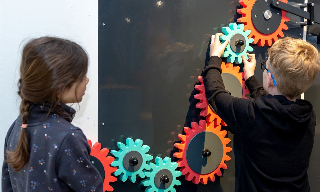 In der Ausstellung bauen Besuchende eine große Kettenreaktion. Schon bei der ersten Herausforderung muss getüftelt werden, um den Impuls vom Zahnrad an der Wand entlang weiterzugeben
