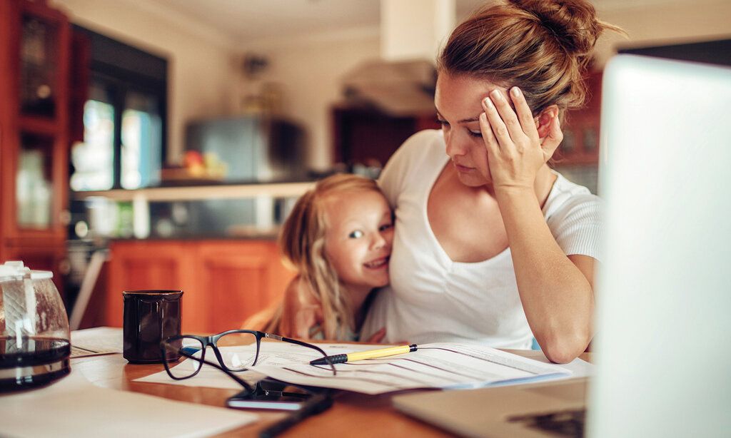 Mutter sitzt am Schreibtisch und ist genervt, kleine Tochter im Arm, die lacht