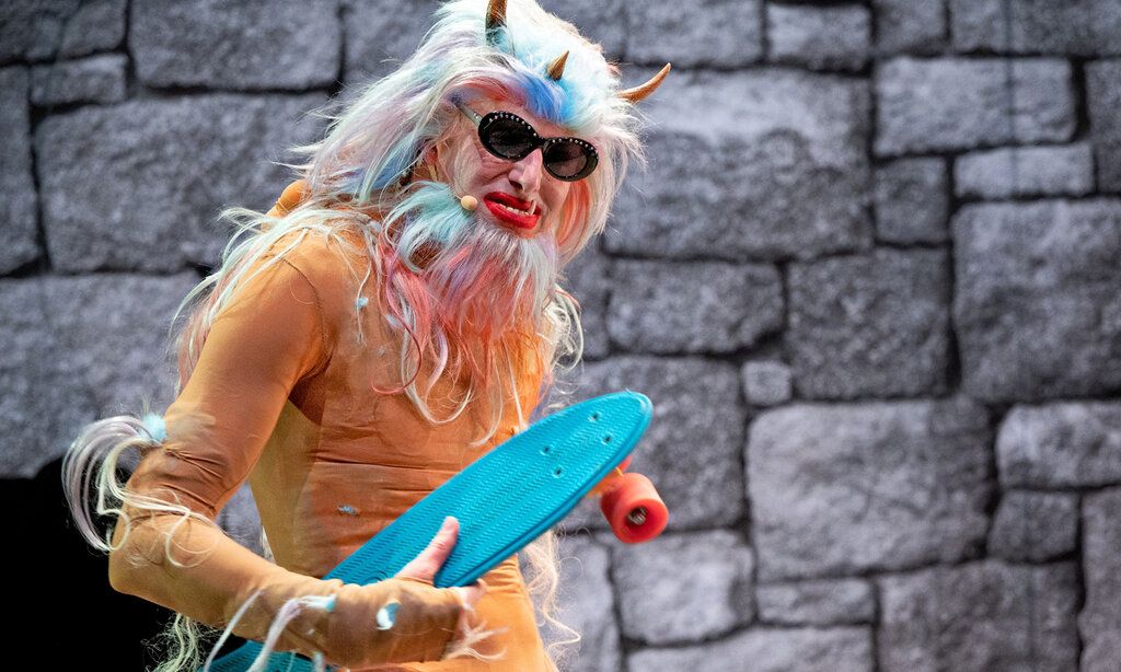 ein als Monster verkleideter Mann steht vor einer Mauer, er trägt ein kleines blaues Skateboard