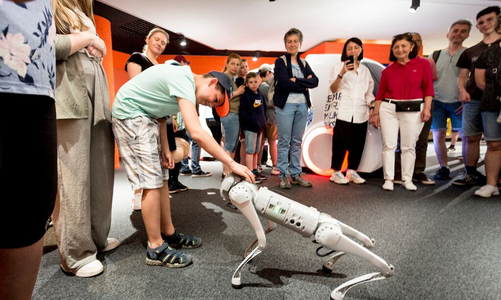 Gruppe von Museumsbesuchern steht zusammen, ein Junge streichelt einen Roboter-Hund
