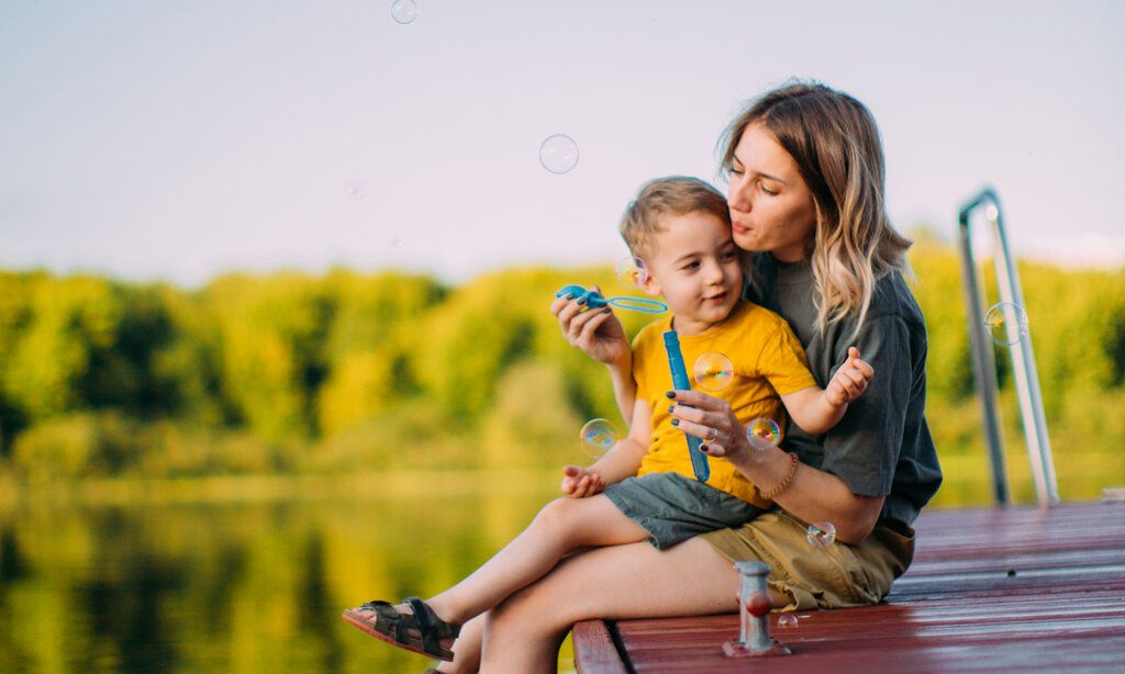 Eine Mutter sitzt mit ihrem Kind auf einem Steg an einem See und pustet Seifenblasen