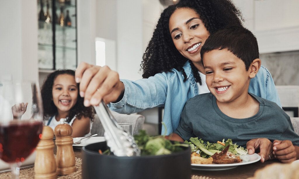 Eine Mutter sitzt mit Tochter und Sohn am Esstisch und tut Salat auf einen Teller