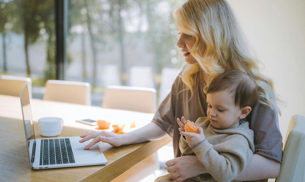 eine Frau guckt auf den Bildschirm des Laptops, auf ihrem Schoß sitzt ein Kleinkind, das eine Mandarine in der Hand hält