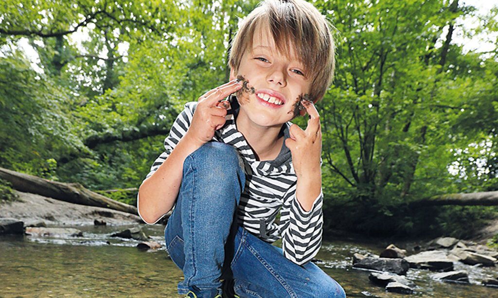 Ein Junge sitzt an einem bach und schmiert sich Matsch ins Gesicht