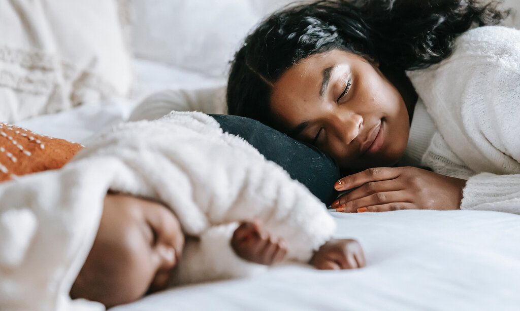 Ein neugeborenes Baby liegt auf einem Bett und schläft, die Mutter schmiegt sich an und schläft auch