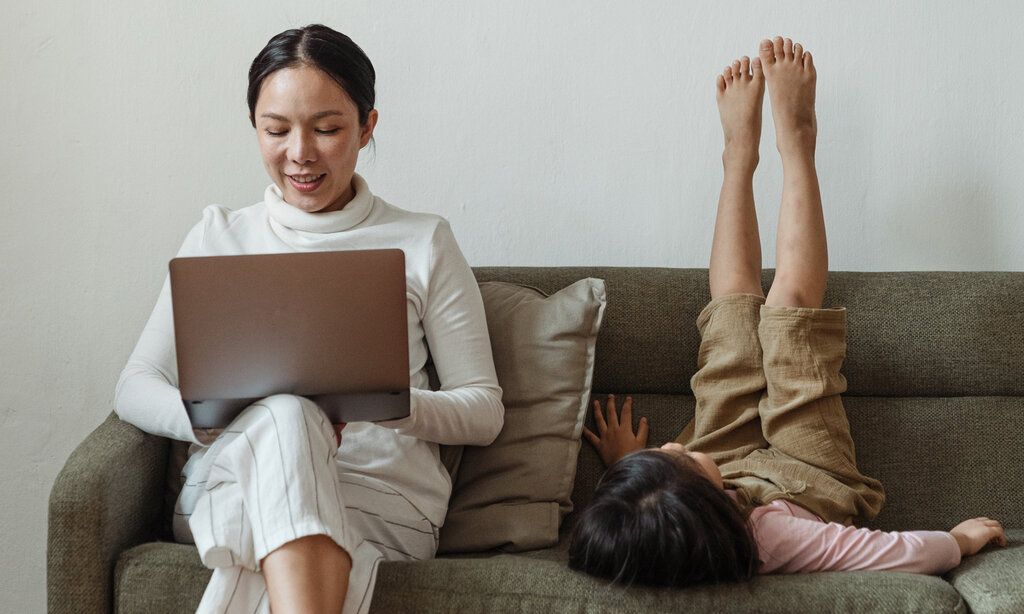 Frau sitzt mit Laptop auf dem Sofa, daneben sitzt ihr Kind verkehrt herum und streckt die Beine in die Höhe
