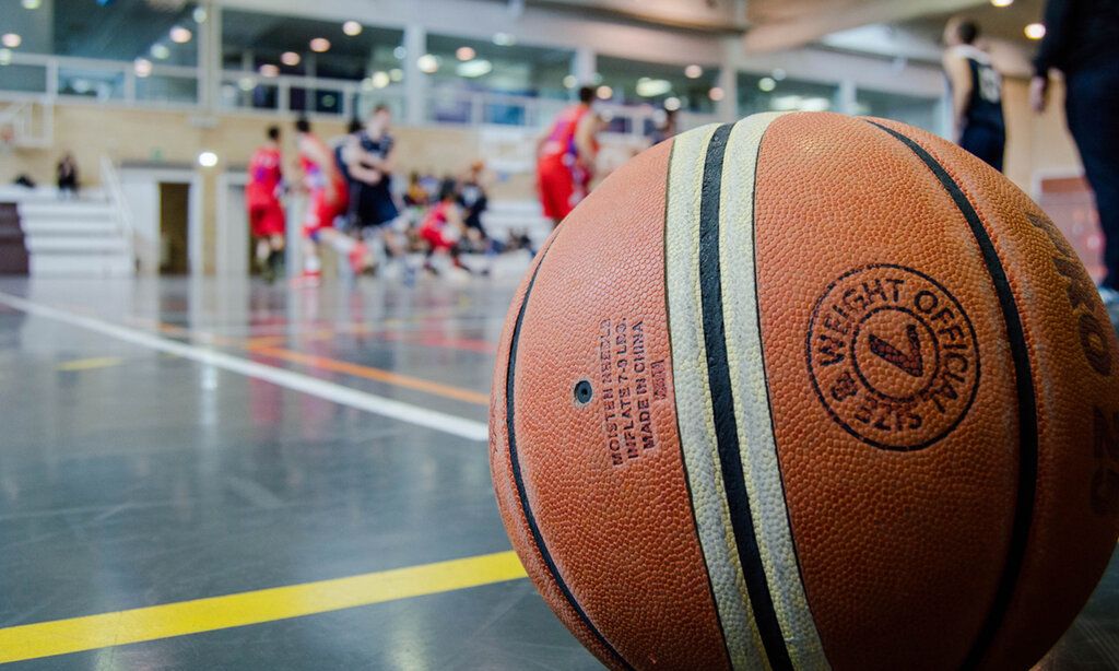 Im Vordergrund ein großer Baskeltball, im Hintergrund eine Sporthalle mit Sportlern