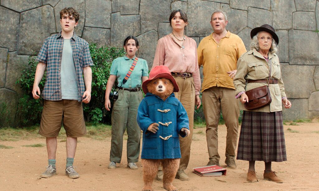 Szene aus dem Film Paddington in Peru, Paddington Bär und fünf Leute (Junge, Mädchen, Frau, Mann, Oma) stehen vor einer Mauer
