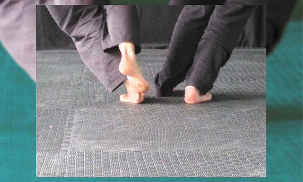 Zwei Menschen, von denen nur die nackten Füsse und die Hosenbeine zu sehen sind, tanzen auf einem Fliesenboden.