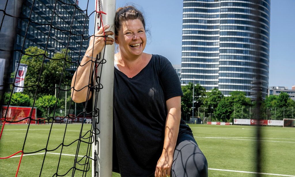 Petra Berg steht auf einem Fußballplatz und lehnt sich an ein Tor. Im Hintergrund Hochhäuser.