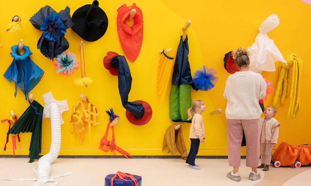 Raum mit gelber Wand und bunten Stoff-Objekten, eine Frau und zwei Kinder stehen zusammen