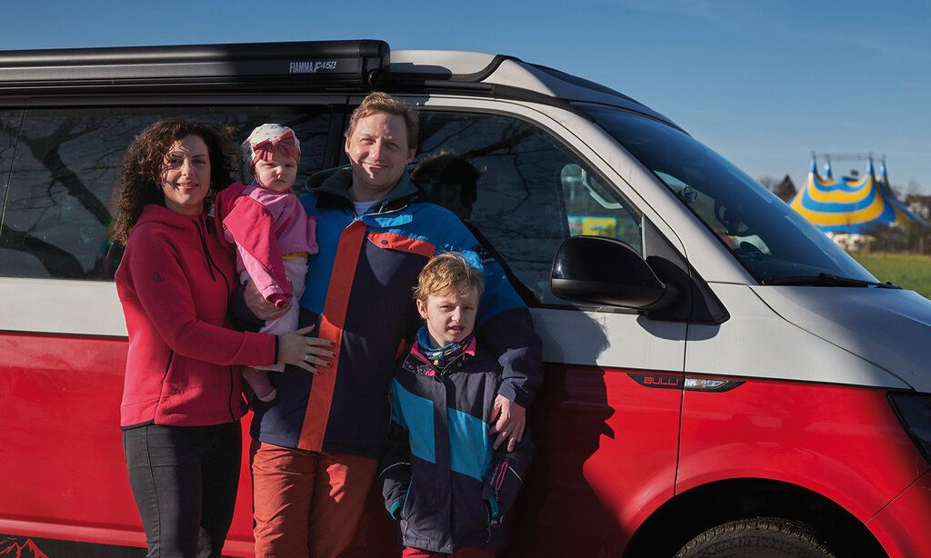 Familie (Vater, Mutter, Kind und Baby) steht vor VW-Bus, im Hintergrung Zirkuszelt, Wintersonne mit harten Schlagschatten