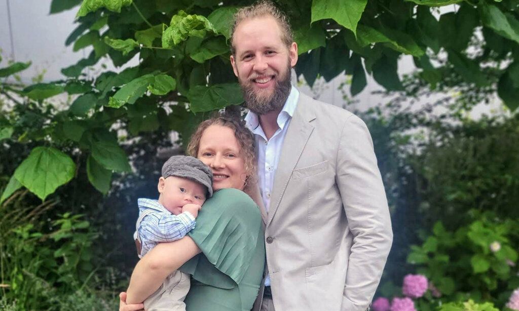 Porträt der Familie Passen, Mutter trägt Baby auf dem Arm, festlich gekleidet, im Hintergund  Pflanzen