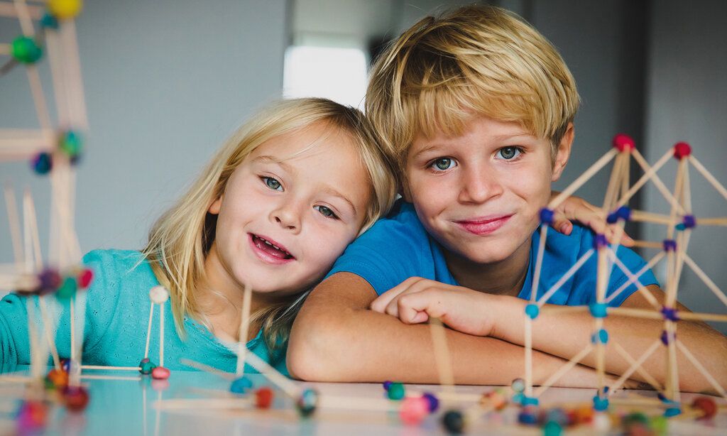 Porträt von zwei Kindern beim Basteln, im Vordergrund unscharf: Gebilde aus Holzstäbchen und Knete