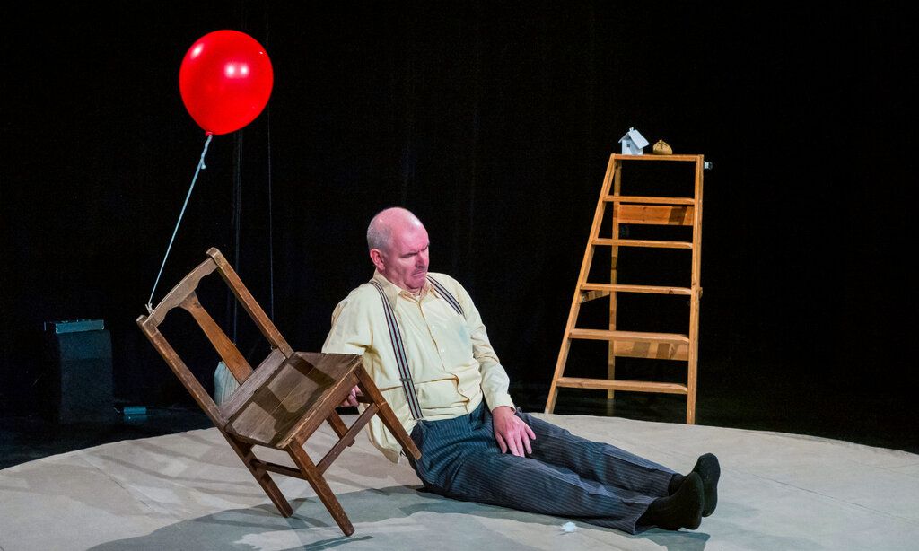 Ein Mann mit Glatze und Hosenträgern sitzt auf einer Bühne zwischen einem Stuhl und einer Holzleiter.