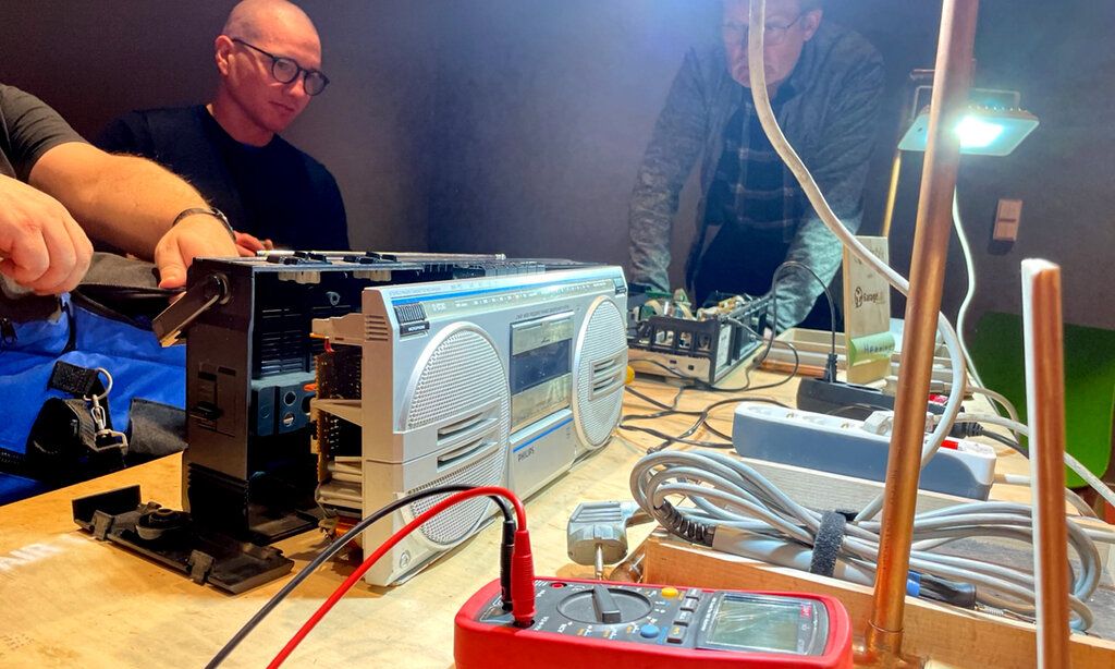 aufgeschraubter Ghettoblaster, Kabel  und Gerätschaften auf einem Tisch, drei Männer beim Reparieren
