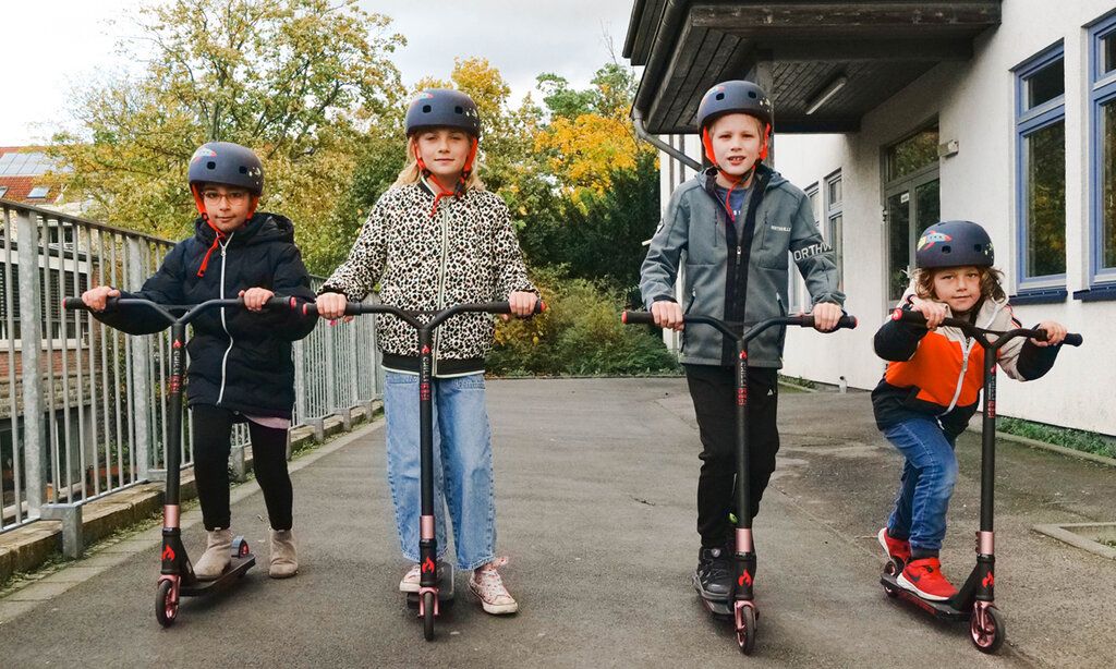 Vier Kinder mit Helmen auf Rollern, schauen in die Kamera