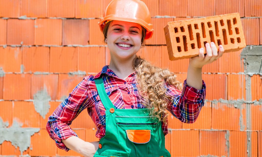 Mädchen mit Bauhelm vor einer gemauerten Wand auf einer Baustelle.