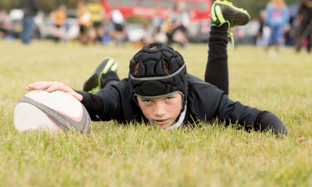 ein junger Rugbyspieler liegt auf dem Rasenplatz, guckt in die Kamera und hält mit einer Hand den Ball fest