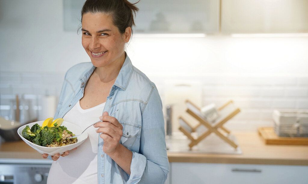 Schwangere Frau steht in einer Küche, lacht in die Kamera und hält einen Teller in der Hand mit Brokoli