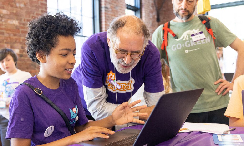 Junge mit violettem T-Shirt arbeitet an einem Laptop, ein älterer Mann im violetten T-Shirt steht neben ihm und spricht mit ihm, ein anderen Mann steht daneben und schaut zu