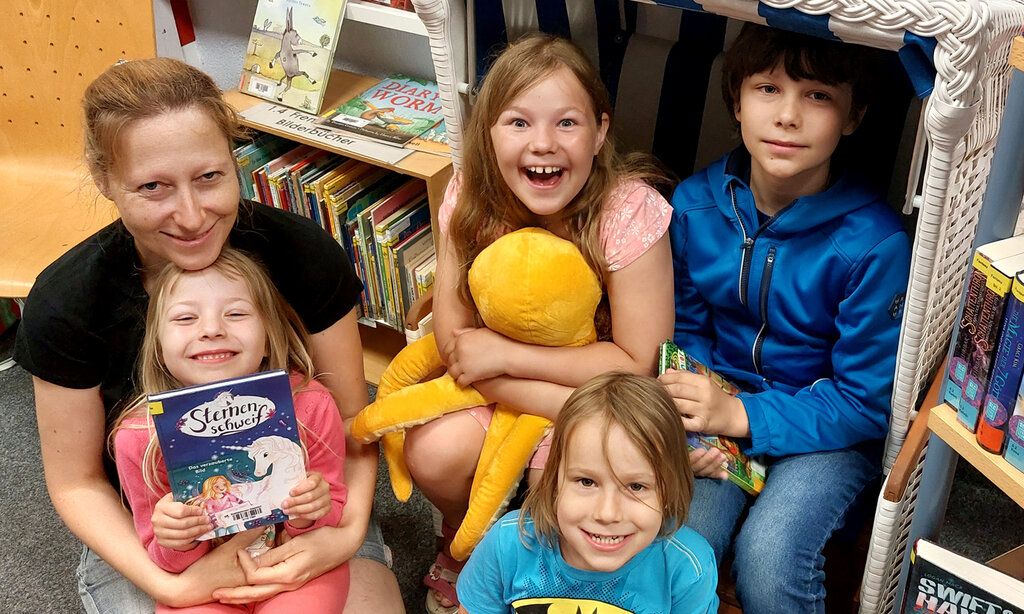 Gruppenbild: eine Frau und vier Kinder sitzen in einer Stadtbücherei auf dem Boden, ein Kind hat ein Buch in der Hand, ein anderes ein Stofftier