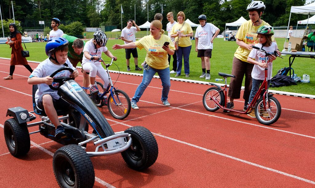 Kinder mit sonderpädagogischem Unterstützungsbedarf fahren mit verschiedenen Fahrzeugen auf einer Tartanbahn, Erwachsene gucken zu und helfen