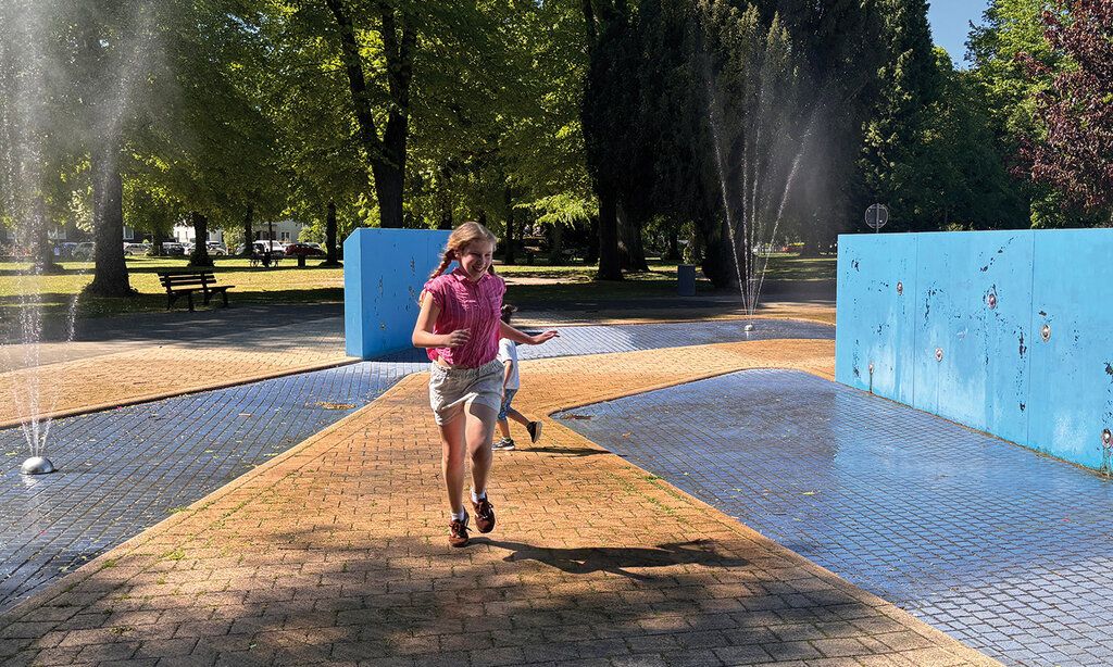 ein Mädchen rennt zwischen Wasserfontänen her auf dem Spielplatz Alter Bilker Friedhof