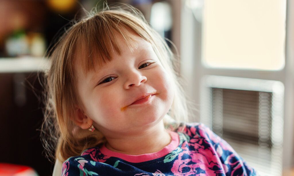 Kleines Mädchen guckt frech grinsend in die Kamera, im HIntergrund unscharfer Raum