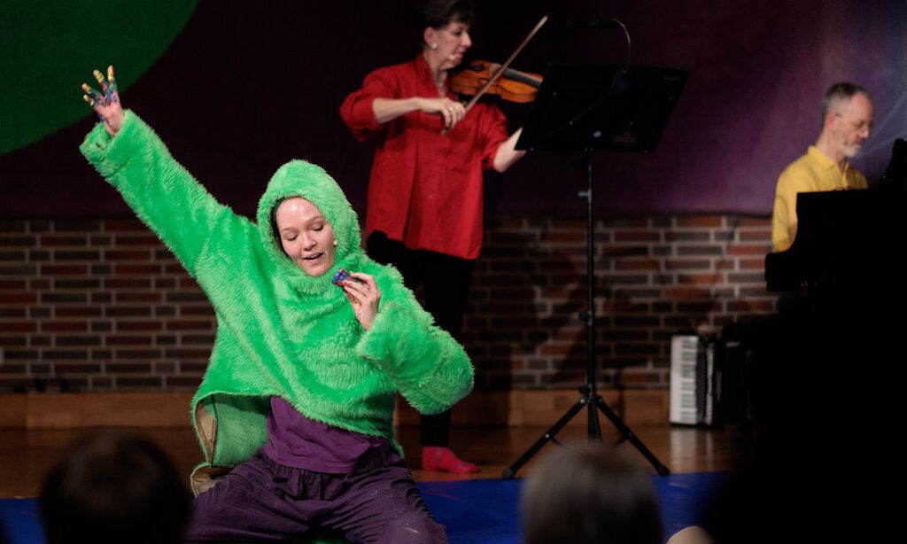 eine Frau mit einem grünen Teddy-Pullover kniet auf der Bühne und streckt den Arm hoch, im Hintergrund: eine Geigenspielerin und ein Mann am Klavier