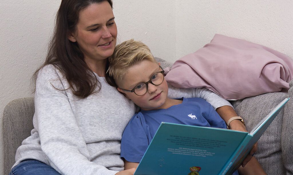 Mutter und Sohn sitzen auf dem Sofa. Das Kind liegt im Arm der Mutter. Die Mutter liest aus einem Buch vor. Perspektive von vorne.